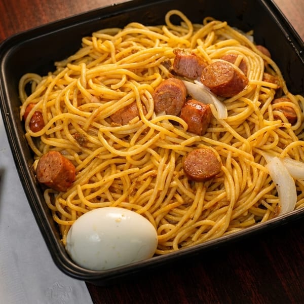 Spaghetti with sausage, onions, and a hard-boiled egg in a black container, with cheese on a napkin.