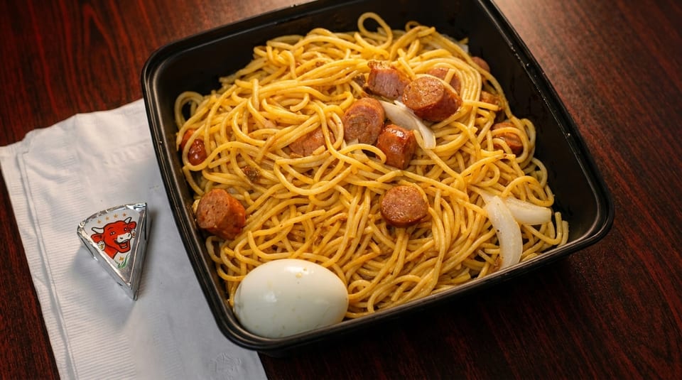 Spaghetti with sausage, onions, and a hard-boiled egg in a black container, with cheese on a napkin.