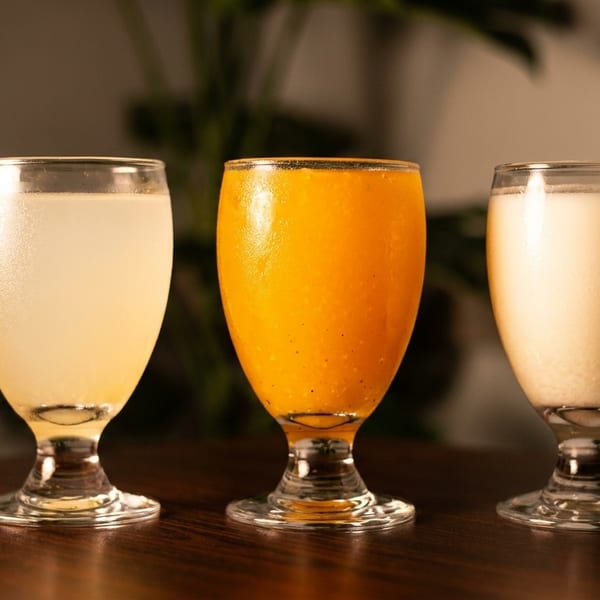Three glasses of different colored beverages, including light yellow, orange, and creamy white.