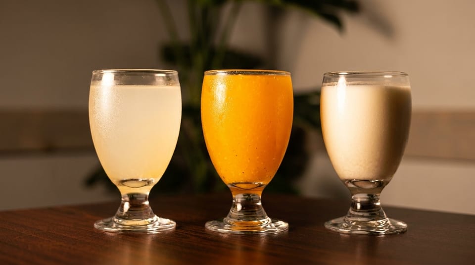 Three glasses of different colored beverages, including light yellow, orange, and creamy white.
