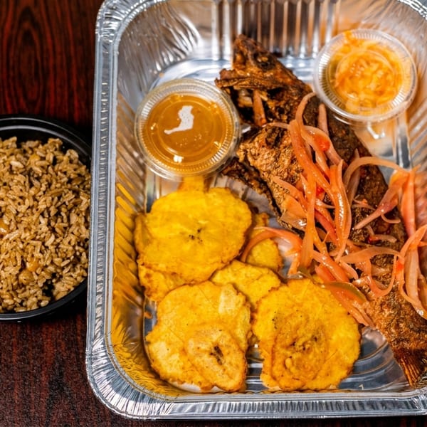 Fried fish with pickled onions, plantains, and sauces in a foil container, with a side of rice.