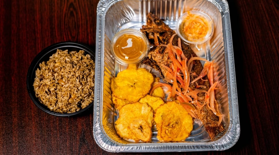 Fried fish with pickled onions, plantains, and sauces in a foil container, with a side of rice.